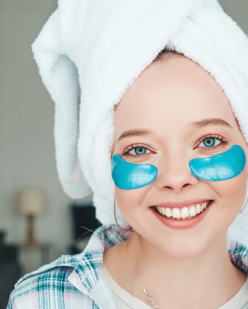 Young beautiful smiling woman with patches under eyes.Sexy carefree model posing in posh apartment or hotel room in white interior.She doing beauty treatments at home in towel on head